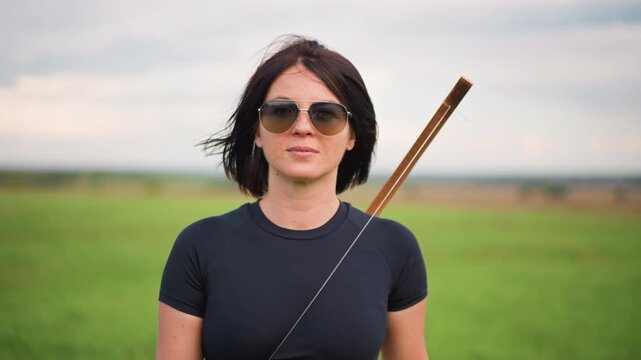 white woman archer in green field, sunglasses, leather quiver over shoulder, holding longbow, windblown hair, calm confident stance, moody sky, cinematic shallow depth, portrait framing
