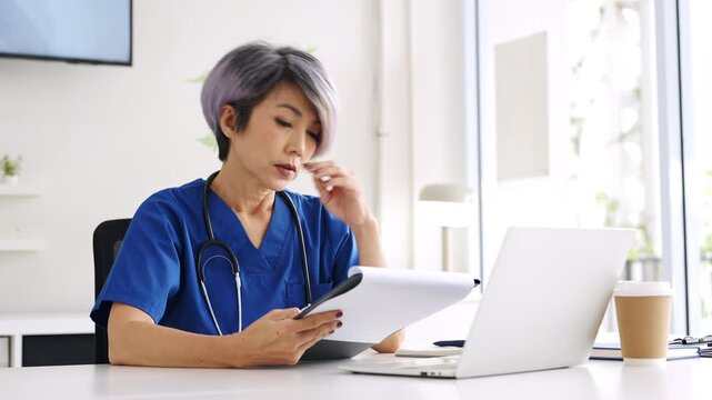 Healthcare worker in scrubs sits at desk, holding a  patient medical charts , rubbing her temple and showing fatigue, stress and burnout from medical frontline pressures