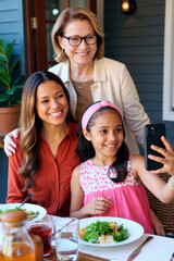 Caucasian middle aged woman standing behind young adult Caucasian woman and Caucasian girl sitting at table smiling while girl taking selfie with smartphone during meal outdoors