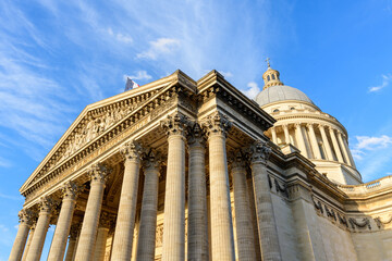 Upward view of the Pantheon's grand neoclassical facade in Paris, featuring ornate columns and a sculpted pediment illuminated by bright afternoon sunlight against a vivid blue sky.