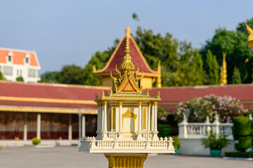 A decorative spirit house with ornate golden Khmer details stands on a pedestal in the open courtyard of the Royal Palace, Phnom Penh, Cambodia. Sunlight highlights the intricate architectural