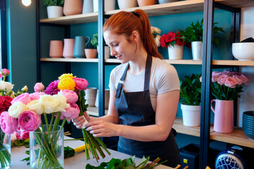 Young adult Caucasian woman arranging colorful flower bouquets in modern floral shop, standing at worktable with various fresh flowers and plants visible on shelves behind her