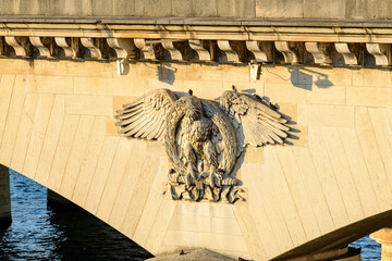 Detailed stone carving of an eagle with outstretched wings decorates the side of Pont de Iena in Paris, illuminated by warm golden sunlight above the flowing river.