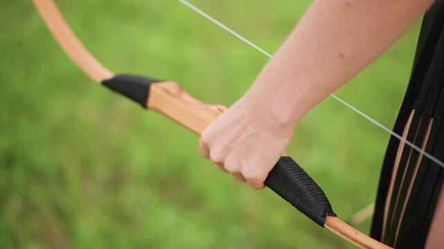 closeup caucasian female hand drawing bowstring, lush green meadow background, visible forearm tension, leather grip, poised release motion, training and empowerment vibe, sunlight on skin, focused