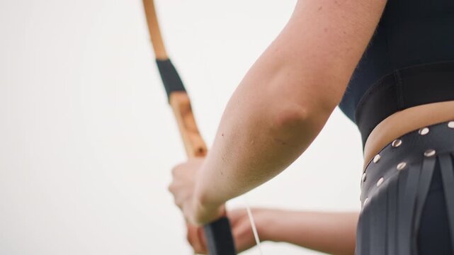 Woman archer drawing wooden bow closeup, leather skirt with studs, steady grip on wrapped handle, arrow nocked, taut bowstring, focused stance against pale sky, cinematic solitary mood