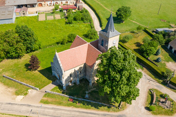 High-angle drone image of a stone church with red tiled roof and a clock tower, surrounded by green fields, trees, and village roads in Cuncy les Varzy under clear sunlight.
