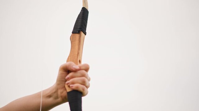 Caucasian archer drawing handcrafted wooden bow, closeup of hand nocking arrow against neutral sky, leatherwrapped grip, taut string, arrowhead detail, focused muscle and steady posture, training
