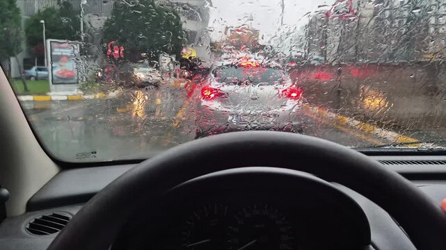 POV inside car during heavy rain, raindrops on windshield with blurred city traffic lights, urban driving in rainy weather conditions.