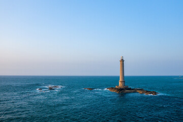 A tall lighthouse stands alone on a small rocky outcrop surrounded by deep blue sea at Cap de la Hague. The scene is illuminated by clear daylight, with gentle waves and a vast open sky creating a
