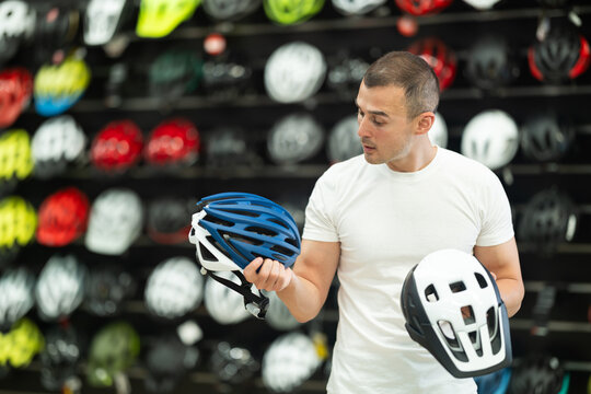 Focused interested male cyclist choosing new bicycle helmet in sports store specializing in cycling gear, comparing design and safety features of two models - Powered by Adobe