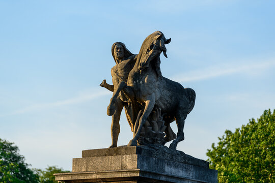 Imposing stone sculpture of a man and horse stands atop a pedestal on Pont de Iena in Paris, highlighted by warm golden sunlight against a clear blue sky and lush green trees.