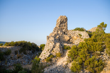 Weathered stone remains of a medieval tower rise above dense green shrubs and rocky ground, illuminated by warm late afternoon sunlight near Chateau de Opoul Perillos.