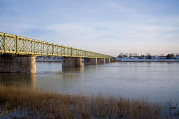 Long yellow iron bridge stretches across the calm Loire River, with snow-covered houses and bare trees lining the distant riverbank under a pale blue sky.