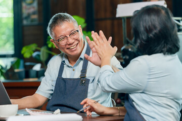 Asian elderly couple cafe owners giving high five and laughing joyfully after success, representing teamwork harmony and partnership in small family coffee business management concept