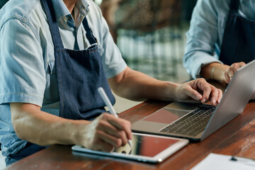 Senior asian male restaurant owner wearing apron using tablet stylus and laptop working on business planning and inventory management inside small cafe workspace during daily operation preparation