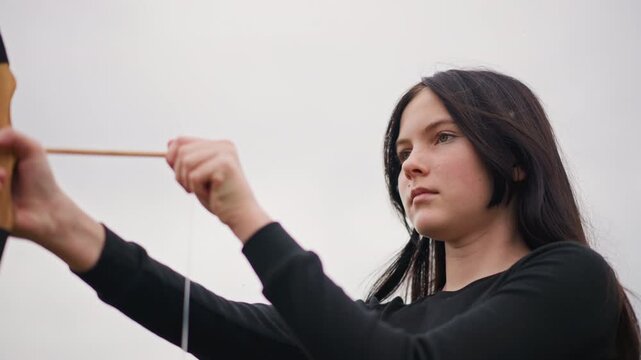 Portrait white woman drawing bow outdoors, eyes closed in calm concentration with braided hair, careful finger placement on string and steady breath, wooden bow curved against overcast sky suggesting