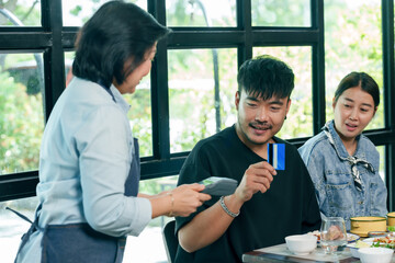 Obraz premium Asian female restaurant owner processes cashless payment with male customer holding credit card while woman watches nearby inside bright cafe scene representing modern small business customer service