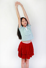 A cheerful young Asian girl in a green striped shirt and red skirt standing and stretching her arms...