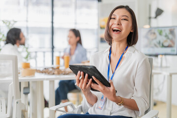 Fototapeta premium Professional Asian businesswoman with short bob haircut smiling excitedly while holding tablet in modern bright office coworking space, blue lanyard, white blouse, happy team meeting background