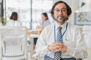 Fototapeta premium Professional Asian businessman with long wavy hair and beard, wearing glasses, white shirt, striped tie and blue lanyard, smiling confidently in modern office coworking space, team meeting background