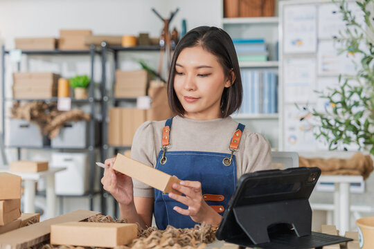 Professional Asian female entrepreneur in beige apron focused on sustainable packaging, checking eco-friendly products on tablet in creative workshop with recycled boxes and olive branches, green  - Powered by Adobe
