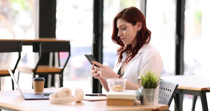 Focused busy caucasian businesswoman in a beige blazer sits at a african ame office desk using her smartphone, checking important information or responding to a message with a concentrated expression
