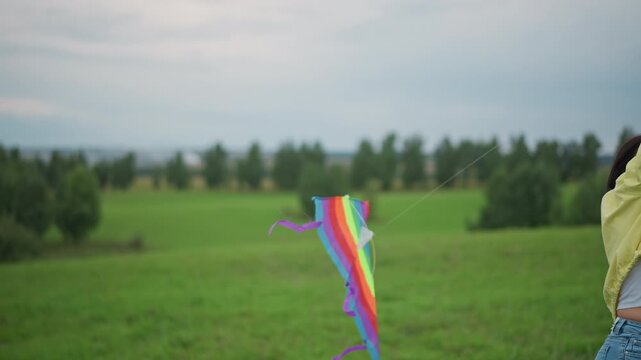 rainbow kite soaring above green field, young woman in yellow jacket runs across meadow, wrist gripping purpletailed kite line, fabric rippling in brisk wind under overcast sky, distant tree line