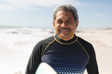 Portrait of smiling retired senior man standing with surfboard at beach on sunny day