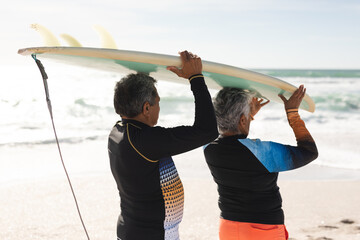 Multiracial senior couple walking together carrying surfboard over heads on shore at sunny beach