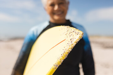 Surfboard with sand held by smiling senior woman at beach during sunny day