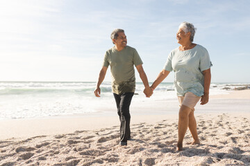 Happy senior multiracial couple holding hands while walking on sand at sunny beach against sky
