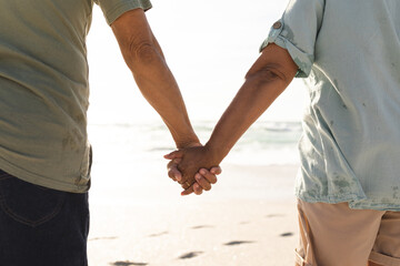 Midsection rear view of senior multiracial couple holding hands at beach on sunny day