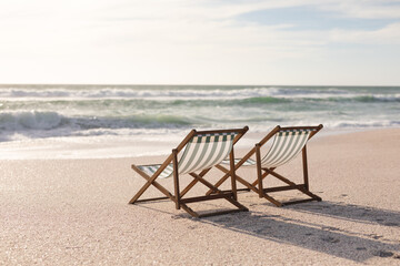 Two empty folding wooden chairs on shore at beach in front of waves splashing in sea against sky