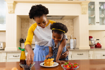 African american mother putting berries over the pancake of her daughter in the kitchen at home