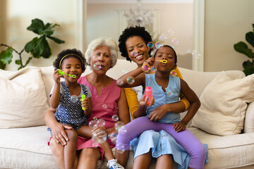 African american grandmother, mother and two granddaughters blowing bubbles sitting on couch