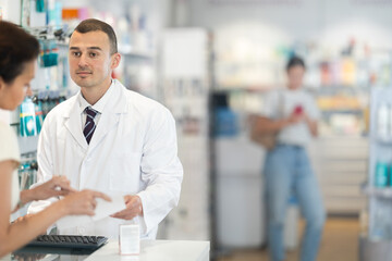 Young male pharmacist giving prescription medicine to adult female buyer at pharmacy