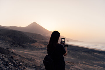 Young Caucasian woman taking photo of Cofete beach at sunset