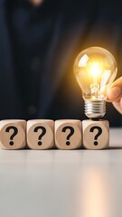 Hand holding a lit light bulb over four wooden cubes with question marks
