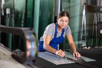 Young workwoman using manual glass cutter to precisely cut sheet of glass in processing workshop environment, showcasing professional skills