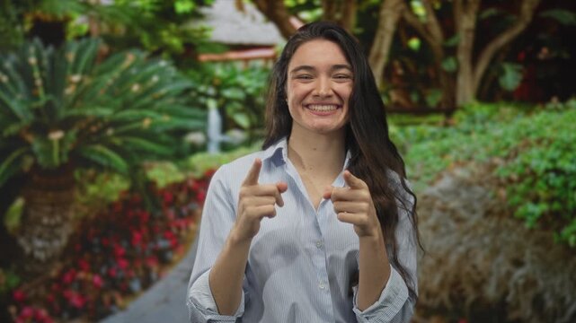 Woman makes ok sign with her hands in green park walkway among lush shrubs and palm trees under dappled sunlight; approval.