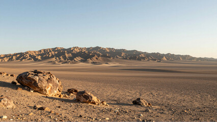 Golden light illuminates a vast, serene desert landscape with scattered ancient rocks and a majestic distant mountain range under a clear sky