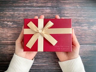 Hands Holding Red Gift Box with Ribbon on Wooden Background
