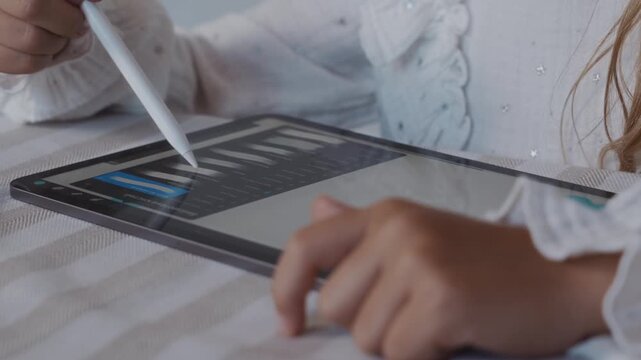 Close up shot from a slight top front angle shows a child selecting tools in a drawing application on a digital tablet using a stylus. The camera emphasizes interface interaction and focused learning.