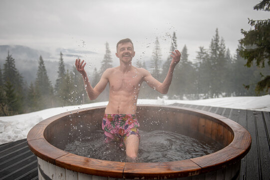 Tub. Guy relax in thermal spa in open air. Man swims in hot bath while resting. Young male enjoying hot tub snowy forest outdoors. Winter holidays in snowy mountains. Hot water treatment concept.