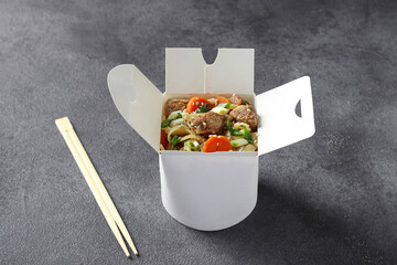 Noodles with pork and vegetables in take-out box on gray table