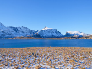 High arctic mountains and steep cliffs rising above deep fjord in winter. Majestic snowy peaks reflecting on calm water surface under bright sunny sky, Lofoten Islands, Norway.