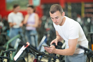 Young guy using smartphone for examining quality seat and choosing bike in modern bicycle sports store