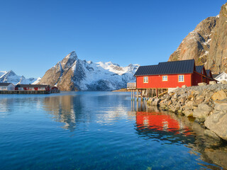 Lofoten Islands fishing village Reine with red rorbu cabins and a pier under a sunny sky in Norway. Crystal reflections on calm water, majestic mountain peaks, and traditional arctic summer scenery.