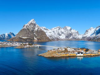 Lofoten Islands fishing village Reine with rorbu cabins, a pier, bridge and islands under a sunny sky in Norway. Crystal reflections on calm water, majestic mountain peaks, arctic summer.