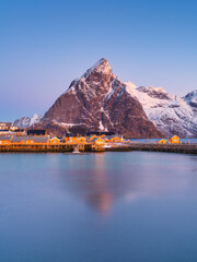 Sunrise over Reine and Hamnoy village, Lofoten Islands, Norway. Iconic red rorbu cabins, snow-covered mountains and fjord illuminated by golden morning light. Scenic arctic winter landscape.